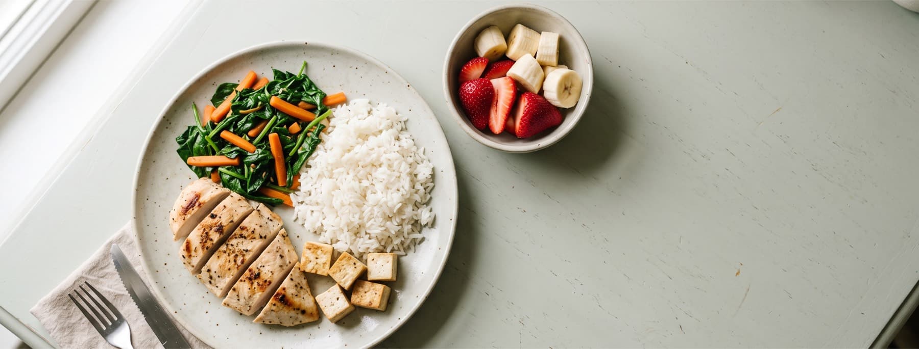 Overhead view of a low-FODMAP plate: sliced grilled chicken, rice, sauteed spinach and carrots, tofu cubes, with a side bowl of strawberries and banana